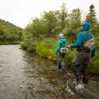 Two people fly fishing in a river surrounded by trees and greenery.