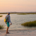 Fly fishing angler wearing a green Fishpond Thunderhead Submersible Sling Pack with orange accents on the beach.